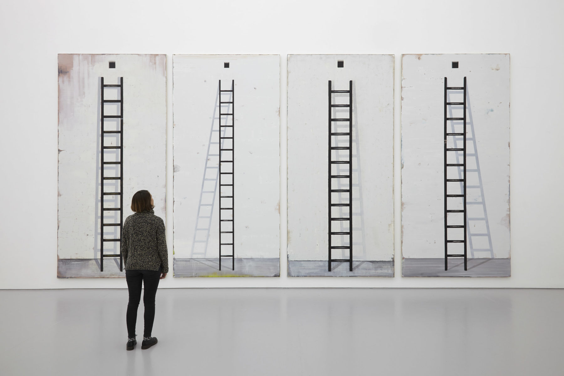 A visitor stands looking at four tall paintings of ladders. Each painting has a shadow appearing differently behind the ladder.