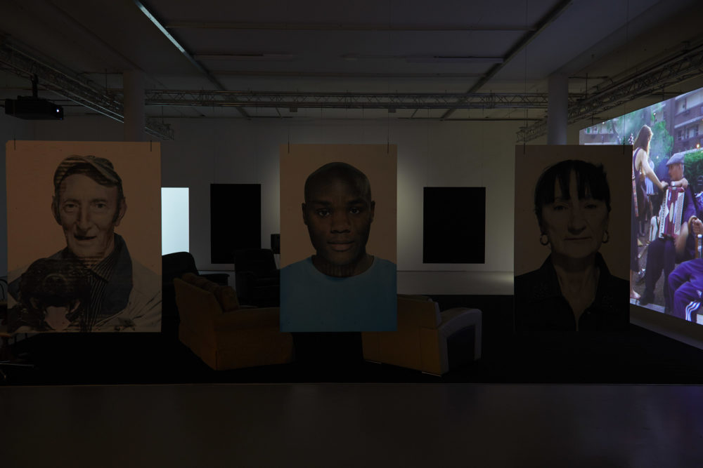 Large portraits of people involved in the project hang from the ceiling, marking a barrier between the gallery walkway and a viewing area with sofas in, pointed at a large screen, currently showing a man playing an accordian.