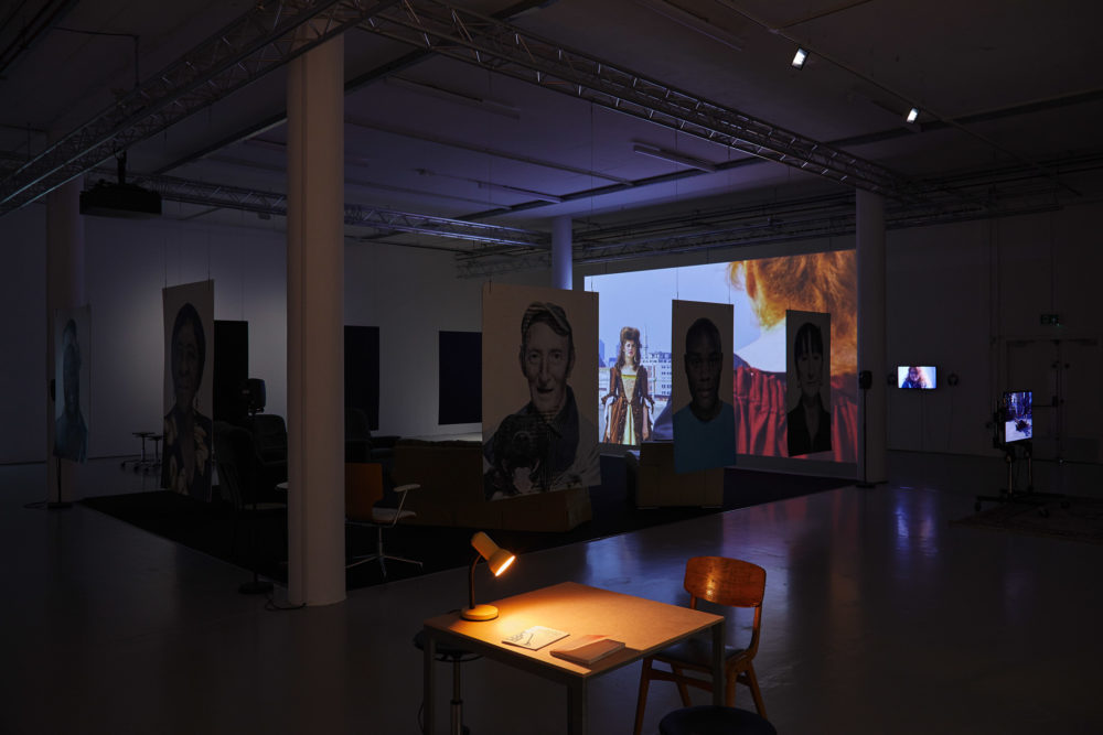 In the foreground a desk is illuminated by an angle poise lamp. Behind the desk are portraits that are hanging from the ceiling.