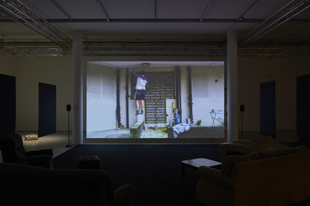 A projection of a man bricking up a doorway hangs in the gallery. Plush sofas and arm chairs point at the projection.