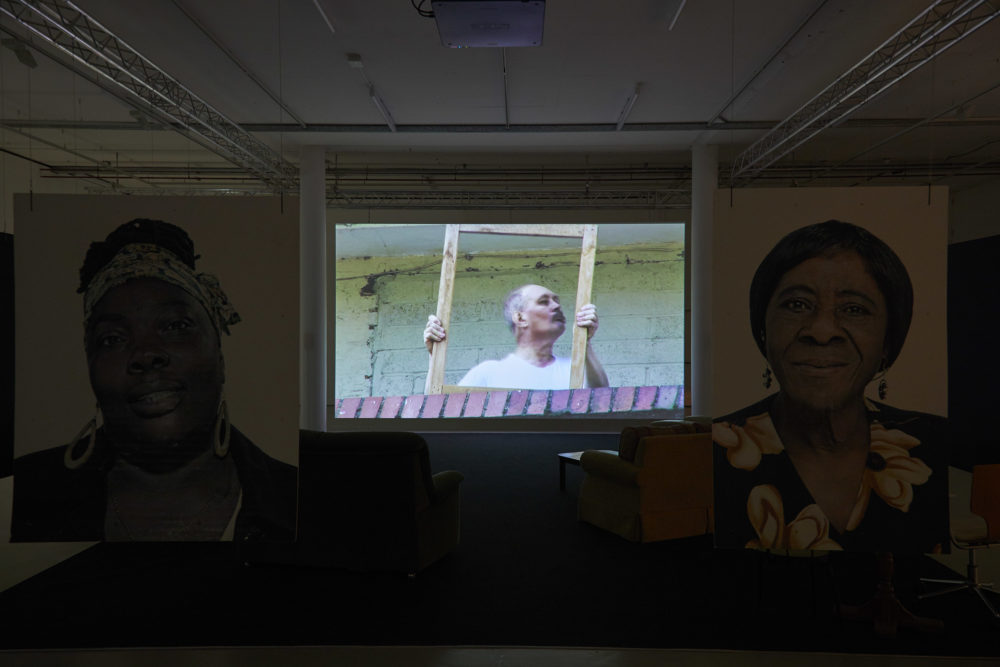 Two large portraits of women involved in the project hang from the ceiling, marking a barrier between the gallery walkway and a viewing area with sofas in, pointed at a large screen, currently showing a man carrying some furniture on a balcony.