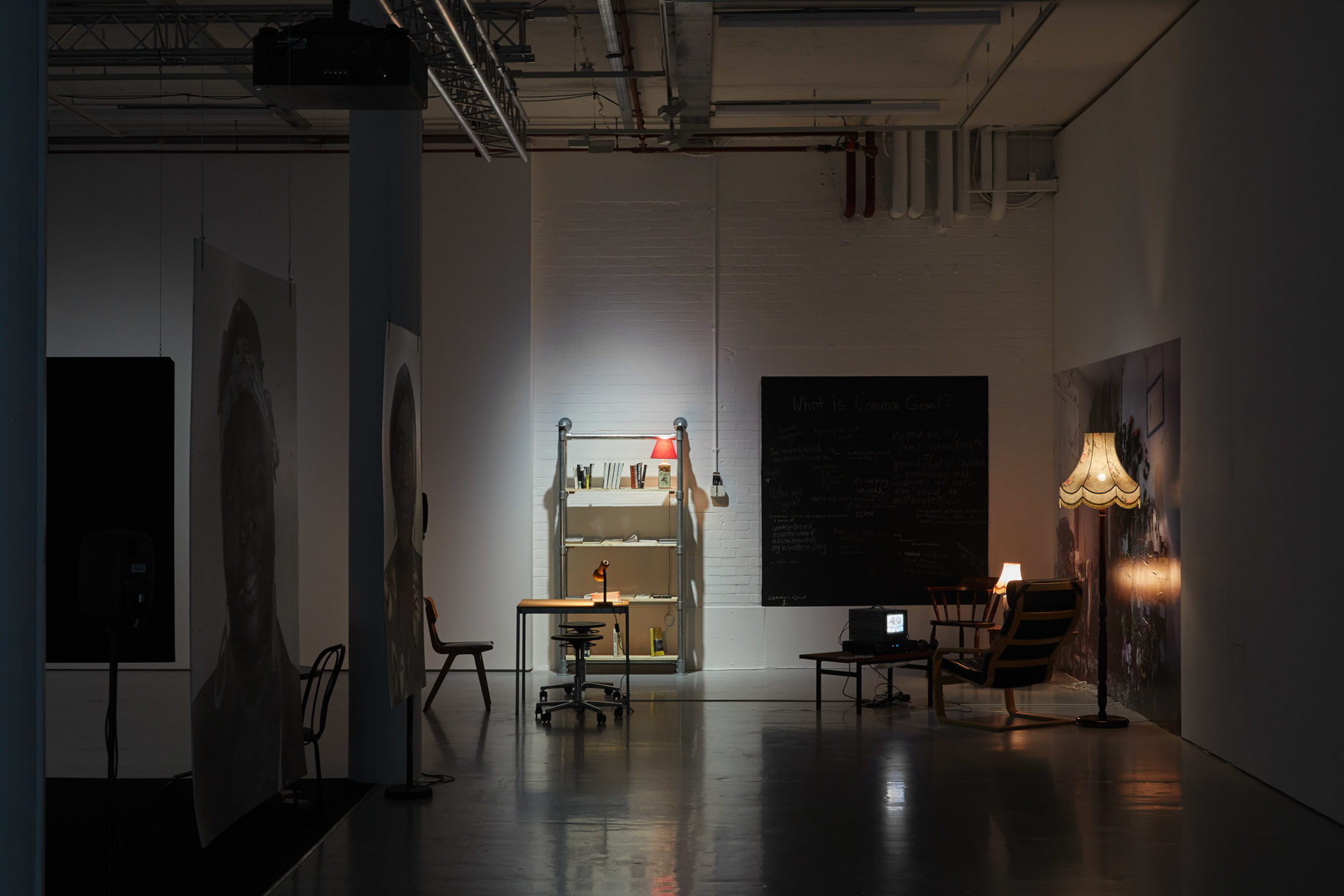 Installation shot: On the far gallery wall is a blackboard, to the left of the blackboard is a book shelf made from scaffolding boards. In the foreground are two large portraits of people involved with the project.