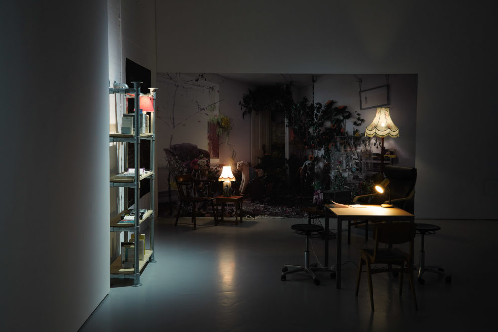 Installation shot: Two tables with lamps, a bookshelf made of scaffolding poles stands in the gallery. On the far wall, a large image of a living room with a lot of plants in it has been pasted to a wall.