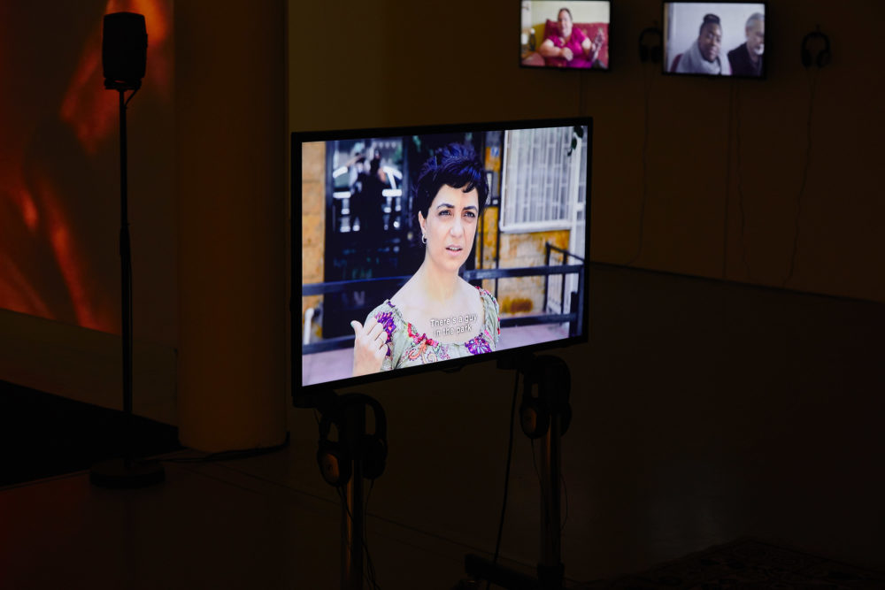 A stand-alone flat screen shows in the gallery, two headphones are positioned just beneath the screen so visitors can hear the audio. On the screen is a woman gesturing behind her and looking confused.