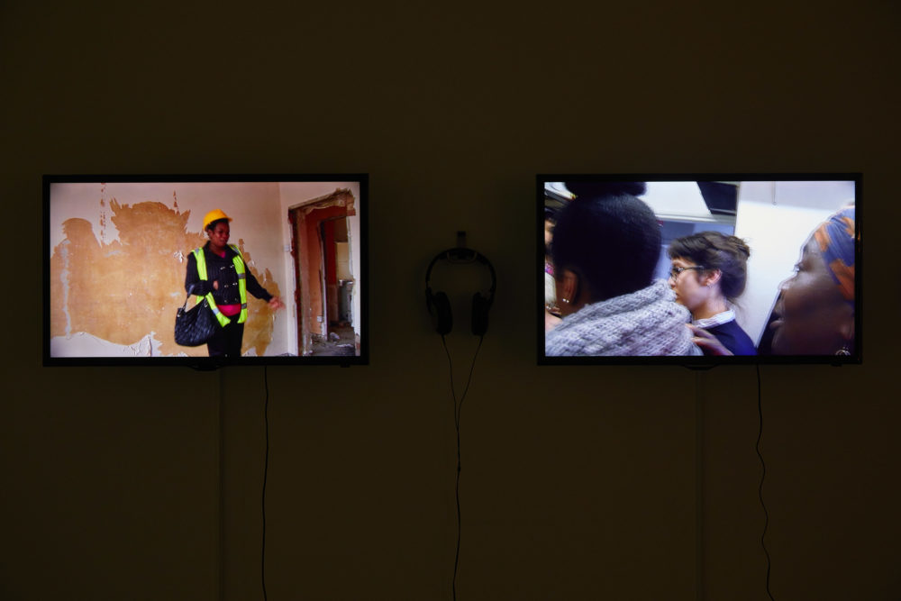 Two flat screen televisions are placed in a gallery - with headphones hung up on a hook between them. The left screen shows a woman wearing a hi-vis vest and points at an empty door frame that looks like its mid construction. The right screen shows three women stood closely with one another - you can only see their shoulders and their heads.