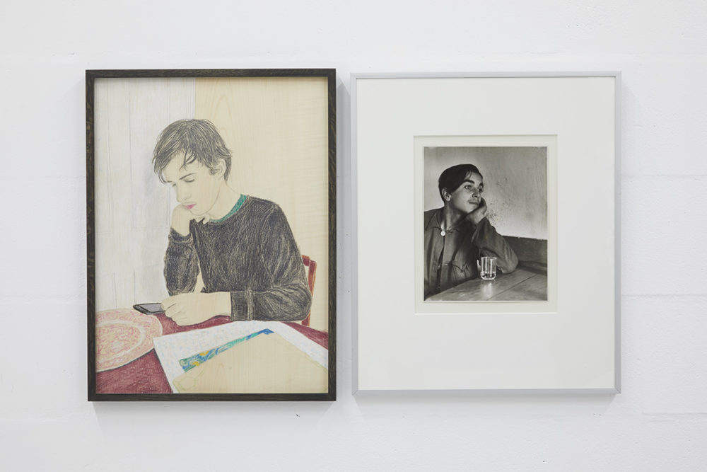 Two framed pictures hang next to one another. On the left, a pencil drawing of a young man sat at his table and looking at his phone. On the right, a photograph of a young person sat at a table with a glass of water.