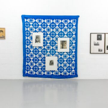 A blue and white patchwork quilt made up of diamond, squares and triangle shapes, hangs on a white gallery wall. Six frames are also hung on that wall - three on top of the patchwork quilt.