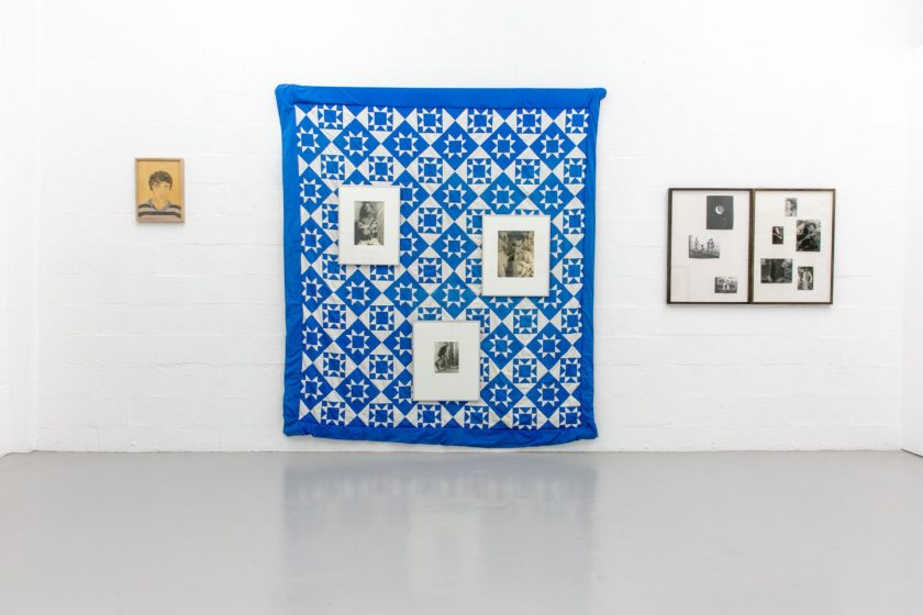 A blue and white patchwork quilt made up of diamond, squares and triangle shapes, hangs on a white gallery wall. Six frames are also hung on that wall - three on top of the patchwork quilt.