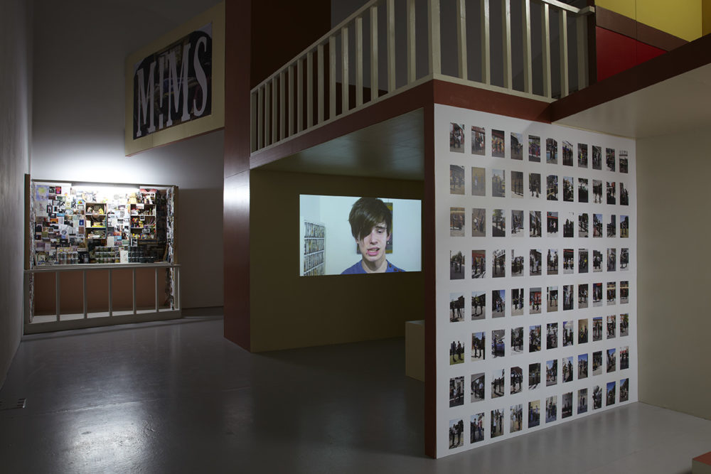 Installation: A dark shelter has been built in the gallery, and an image of a teenage boy looking awkward is projected within it. A balcony appears above the projection.