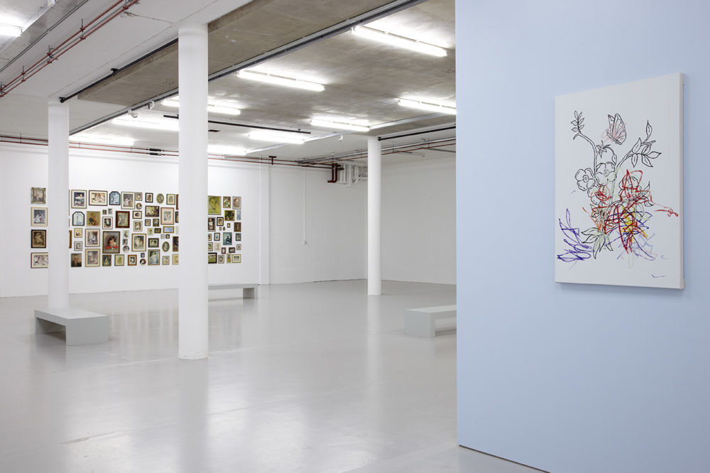 Exhibition: A wall painted cornflower blue has a colouring in canvas picture hung on it. The colouring in picture has been scribbled on with bright crayons.