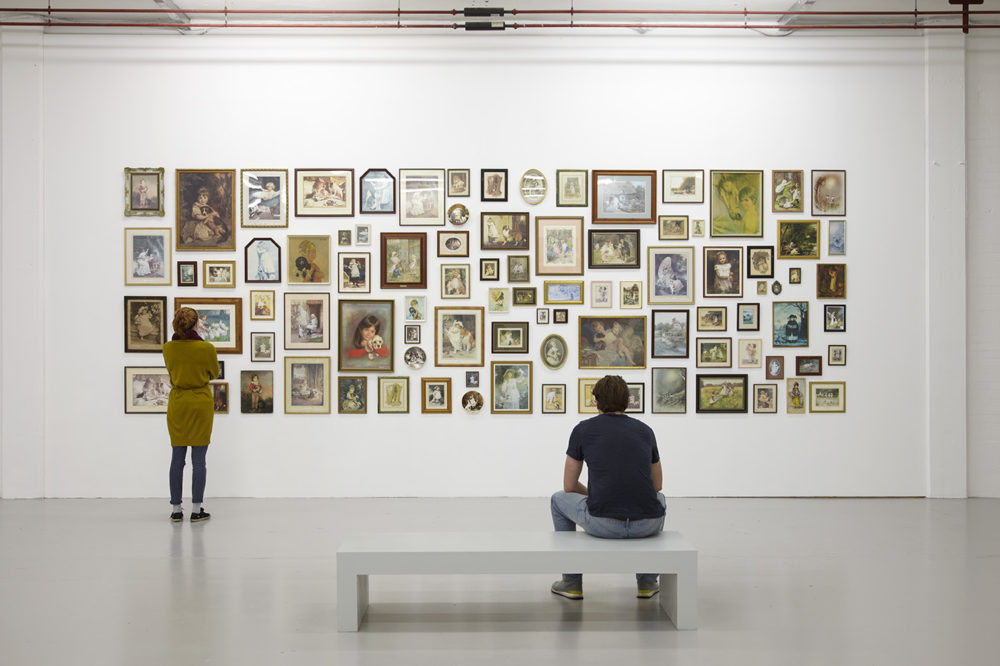 Exhibition: A visitor studies a wall that is hung with so many images in frames that they are clustered together with only an inch or so space between. The images are kitsch, many are drawings of children with pets.