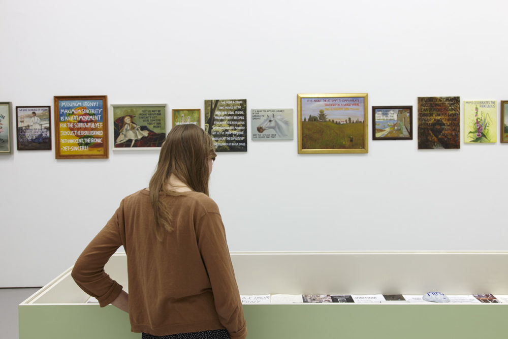 Exhibition: A visitor studies a display case with collected ephemera. Opposite the visitor, on the gallery wall, is hung several images overlaid with text that cannot be read from this viewpoint.