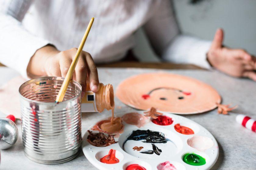 A paint palette is being filled by a young child whose hand is in the foreground and in focus, but whose body is out of focus in the background. They have painted a plate with a smiley face.