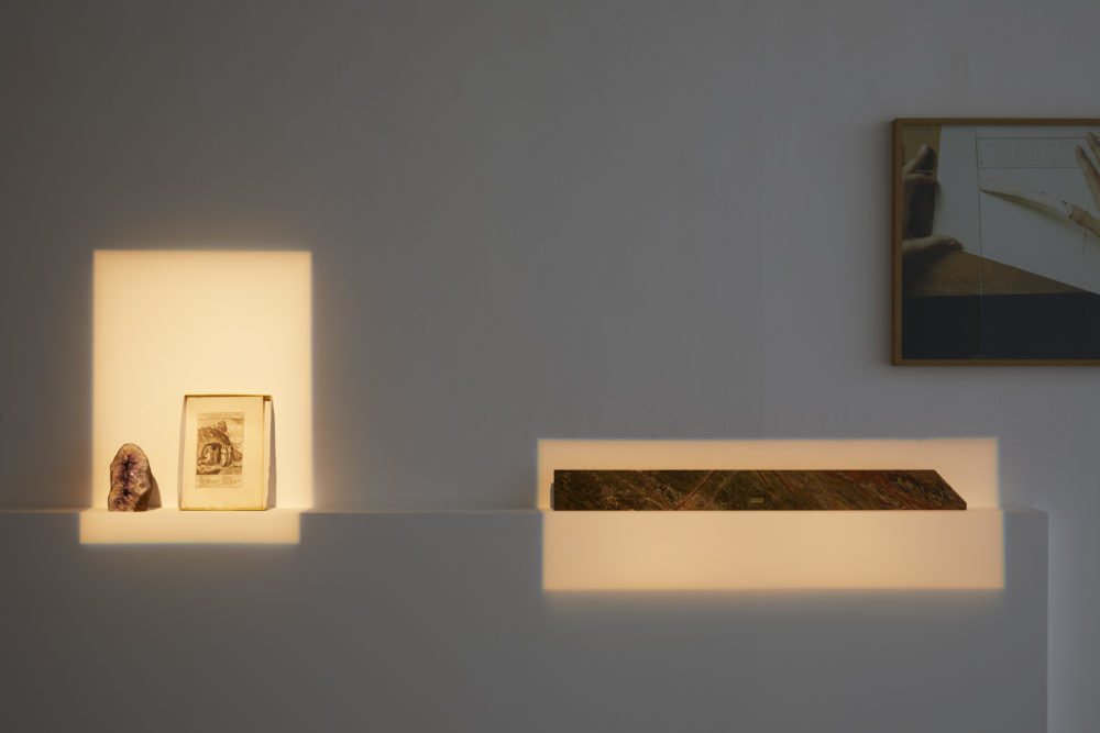 Found items are placed on a shelf in the gallery. The items, left to right are a geode, a page of a book and an oblong of marble.