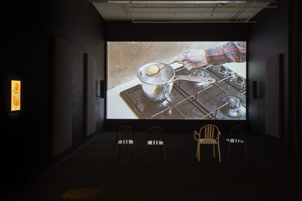 A projection in the gallery shows a pan full of water on a hob and a person getting a boiled egg out of the water using a spoon. There are four empty chairs in front of the screen.