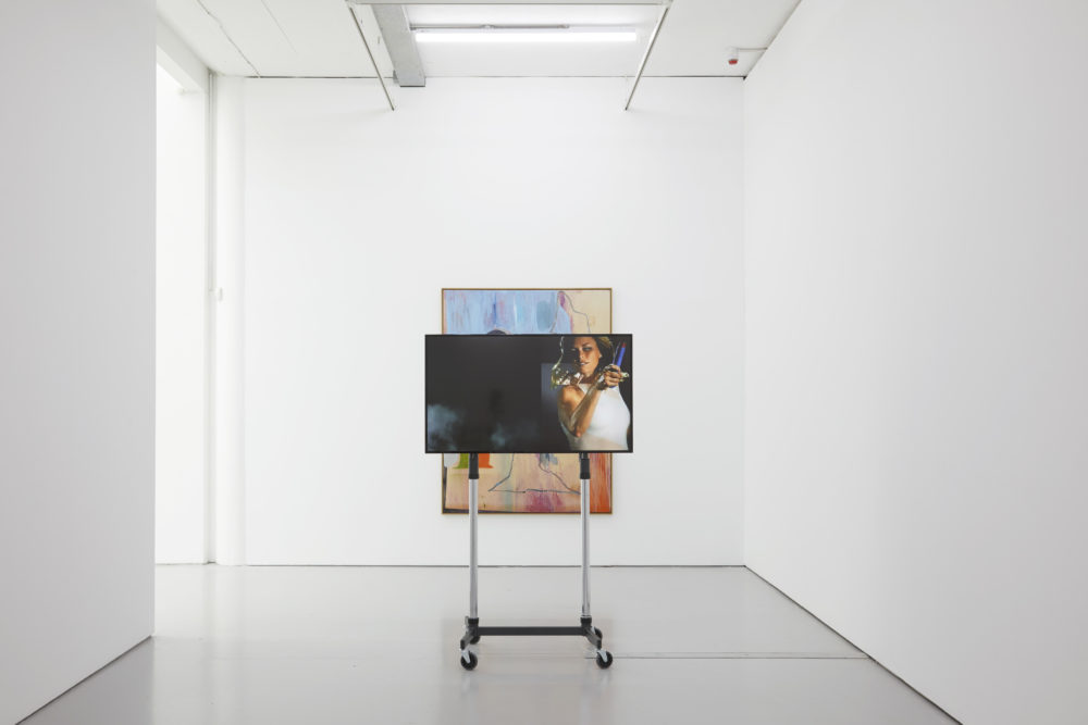 A television screen on a freestanding platform shows a woman in white swinging a tennis rack. Behind the television screen you can see the top and bottom of a painting.