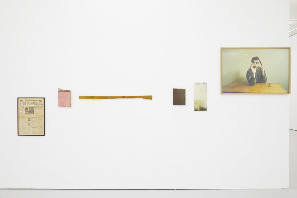 The gallery's white walls have various objects including a newspaper front page and a photograph of a man at a table hung on them.