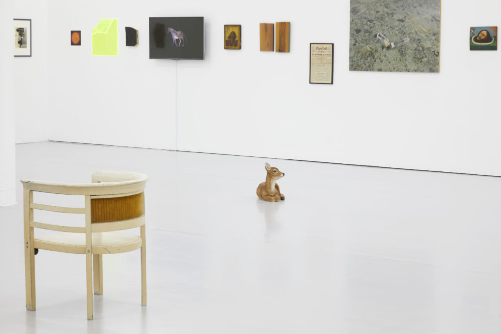 A model of a young deer sits on the floor in the middle of the gallery. In the foreground is a wooden chair. The walls of the gallery are hung with various found paintings, articles and photographs.