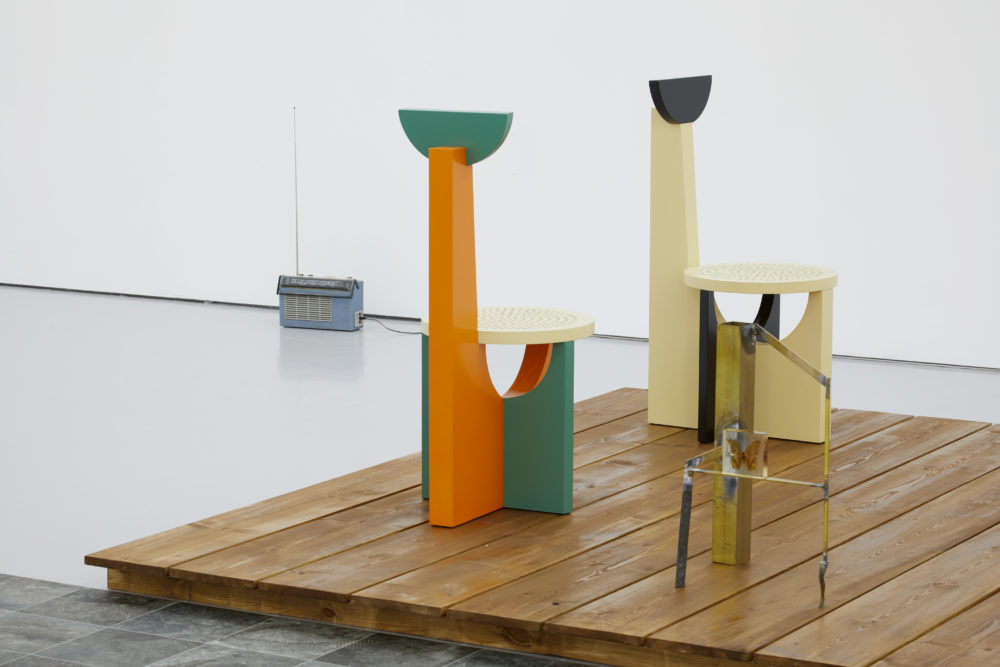 A radio sits on the floor of the perimeter of the gallery, in the foreground is a wooden platform with two brightly coloured, oddly shaped chairs.