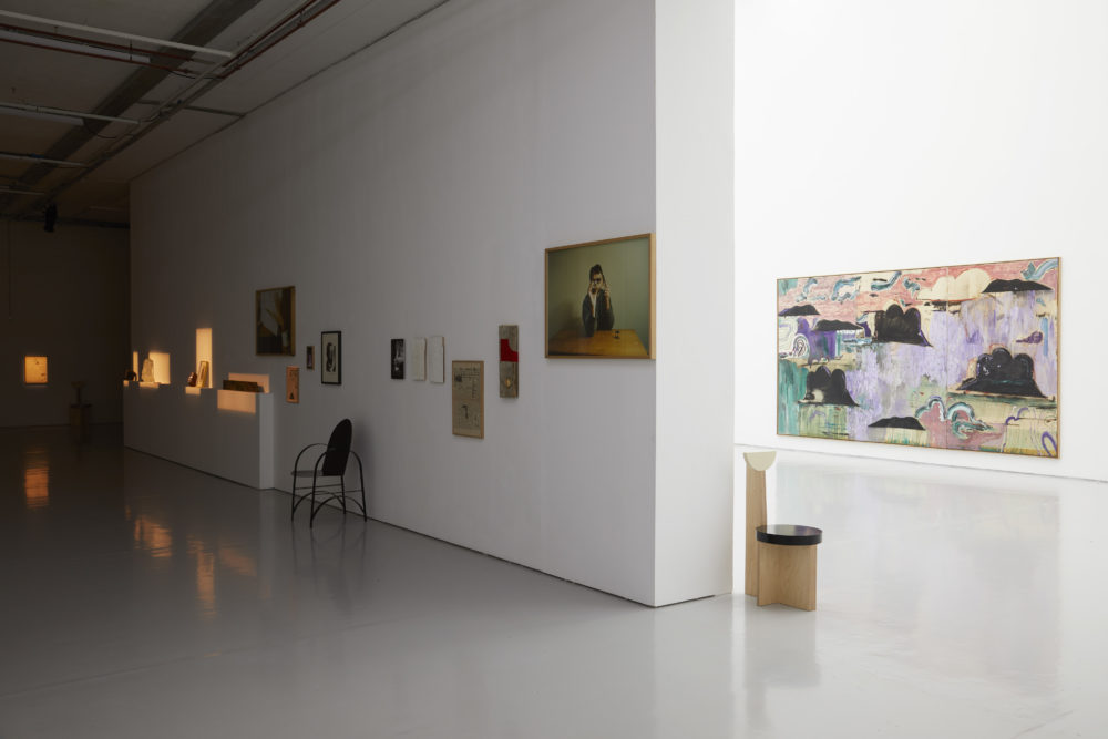 The gallery is divided in two - the corridor that shows to the left of the camera is dimly lit with small pockets of warm light illuminating found objects. The main gallery is brightly lit and an abstract painting of clouds is visible.