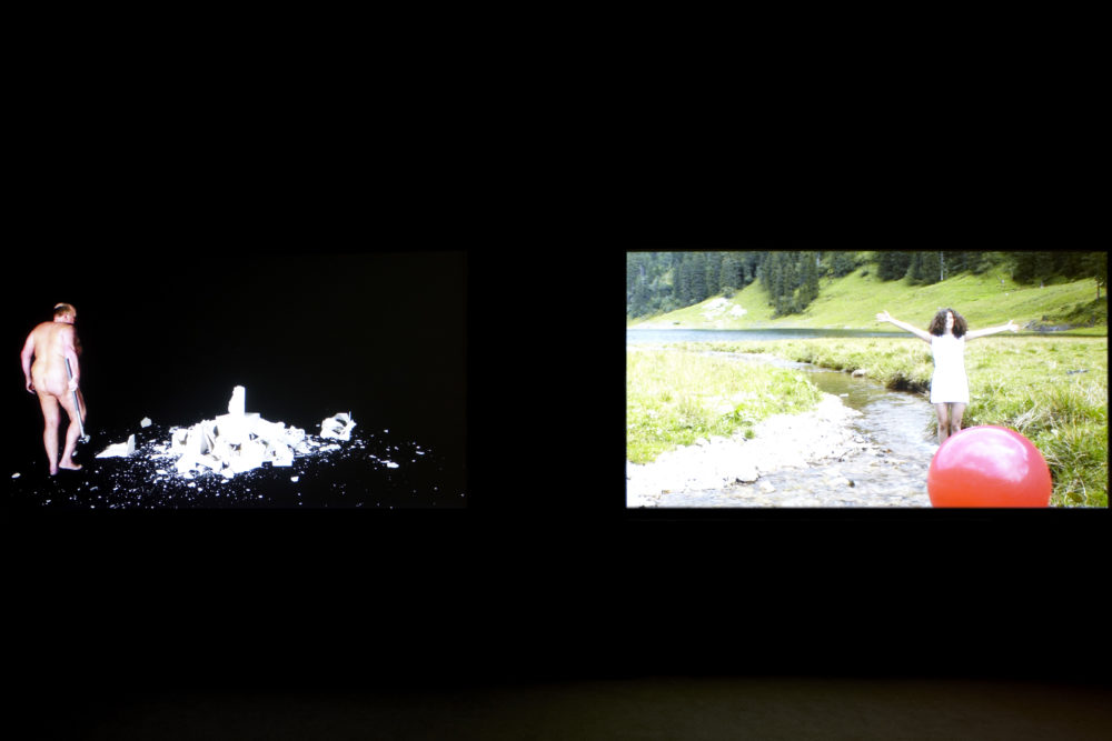 Installation view of Bloomberg New Contemporaries (2013) Two projections in a dark room - one shows a naked man breaking something (now undistinguisable) and another shows a woman in a stream, her arms outstretched and a large red ball in front of her.