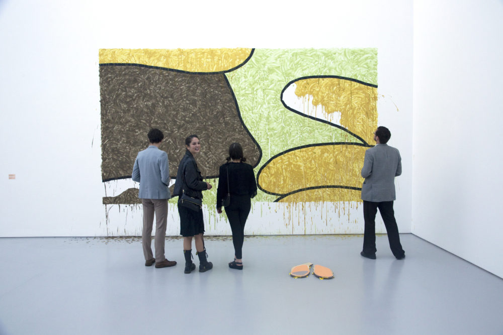 Installation view of Bloomberg New Contemporaries (2013) Visitors study a piece that looks as though it was painted directly on the wall. The image is a close up of Homer Simpson.