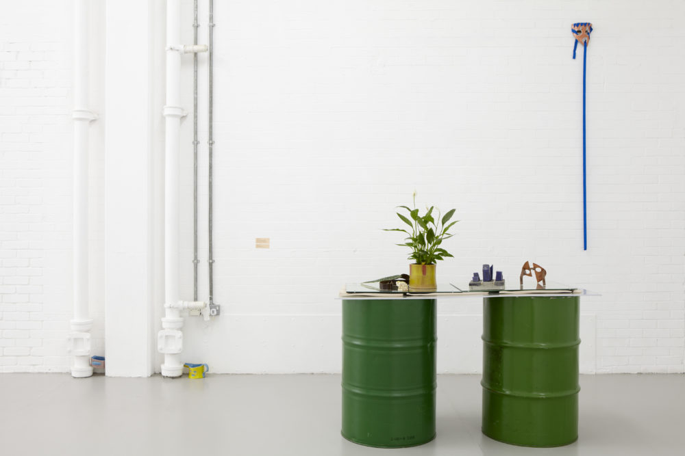 Installation view of Bloomberg New Contemporaries (2013) Two large green barrels have a sheet of perspex draped over them, and a plant pot resting atop that.