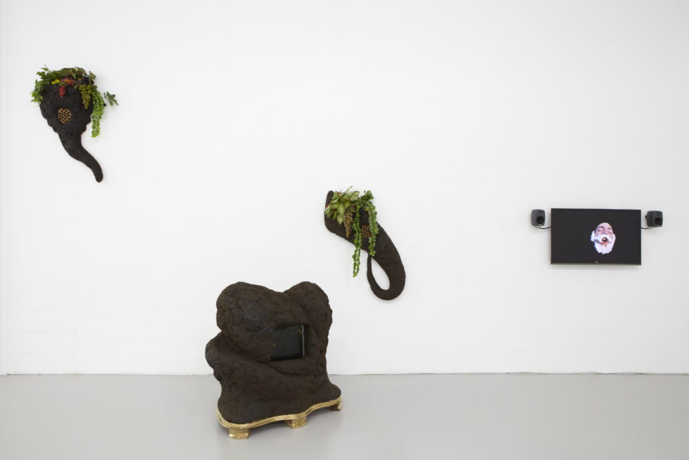 Installation view of Bloomberg New Contemporaries (2013). Sculptures in the gallery appear to be made from mud. A screen shows a person wearing a false beard.