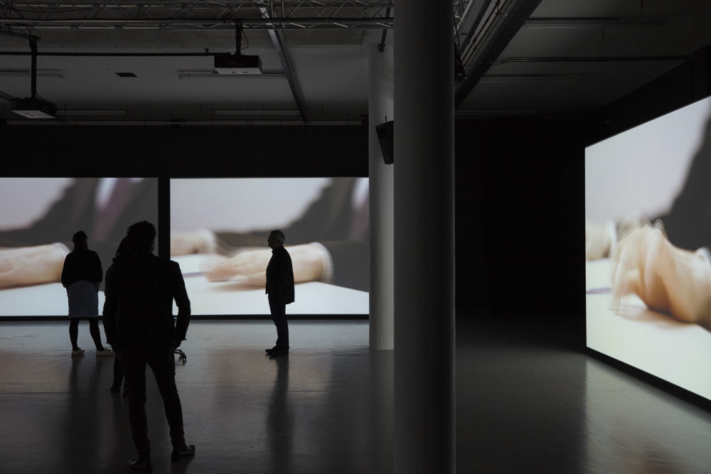 A small group of visitors study the three screens in the gallery. All screens currently show a blurred picture of hands typing on a keyboard.