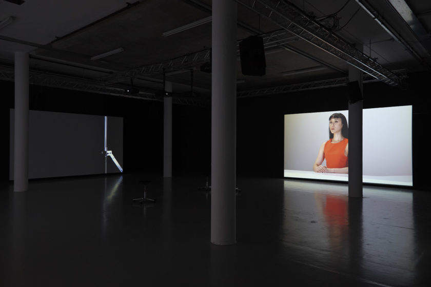 The gallery is dark apart from a large screen showing a woman wearing a red shirt sat with her back straight at a desk. Her facial expression looks stern.