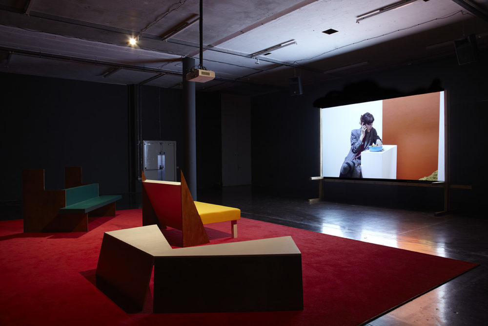 Installation view of Cara Tolmie, Pley (2013). Angular furniture, is illuminated in front of a film. The screen currently shows a seated woman making a phonecall.