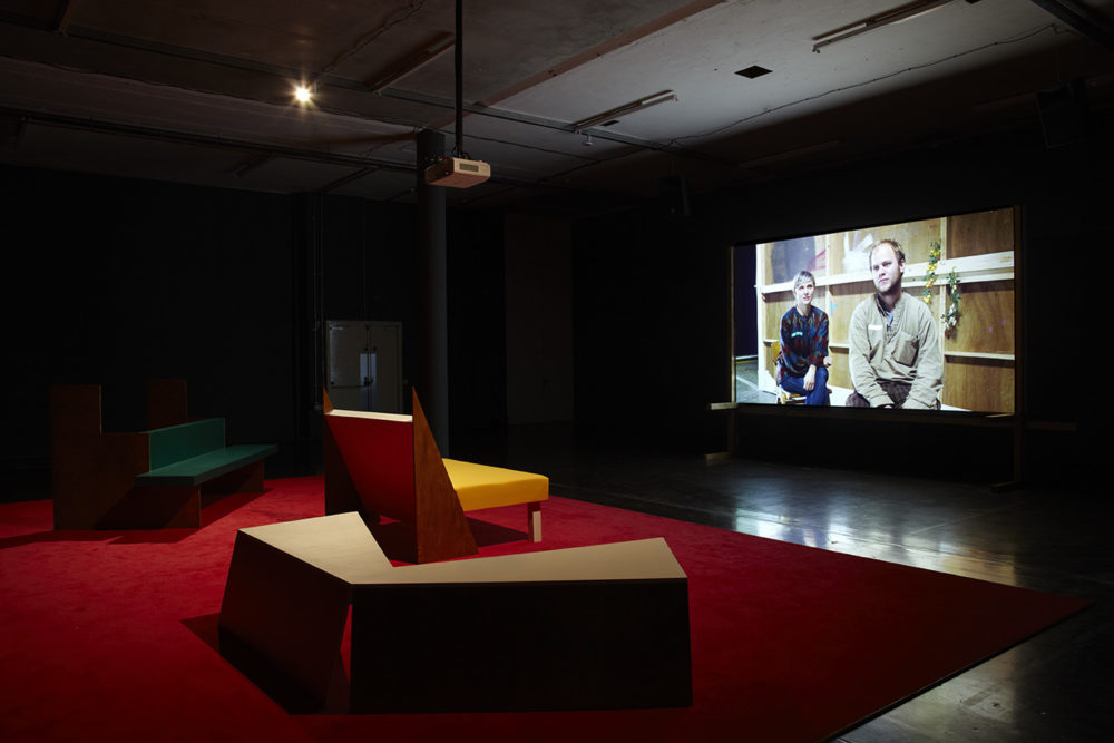 Installation view of Cara Tolmie, Pley (2013). Angular furniture, is illuminated in front of a film. The screen currently shows a man and a woman sitting facing forward.