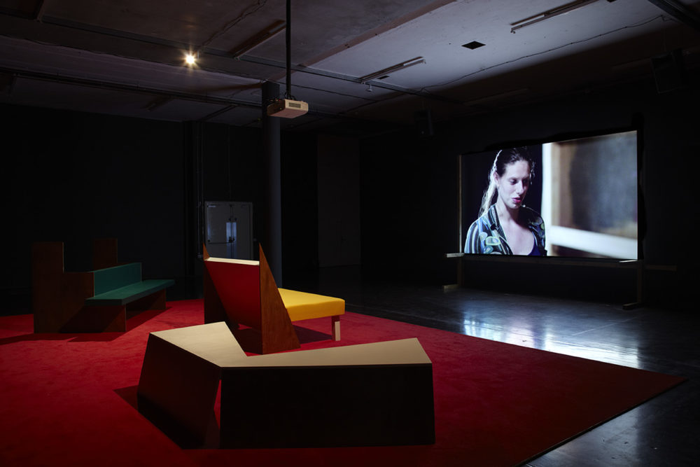 Installation view of Cara Tolmie, Pley (2013). Angular furniture, is illuminated in front of a film. The screen currently shows a close up of a woman.