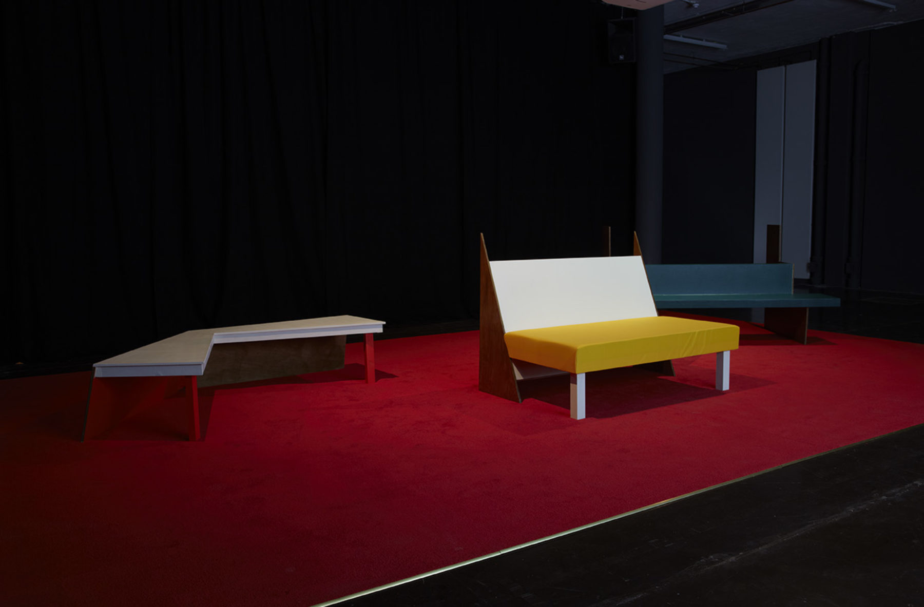 Installation view of Cara Tolmie, Pley (2013). Angular colourful furniture sits spotlighted in a dark room.