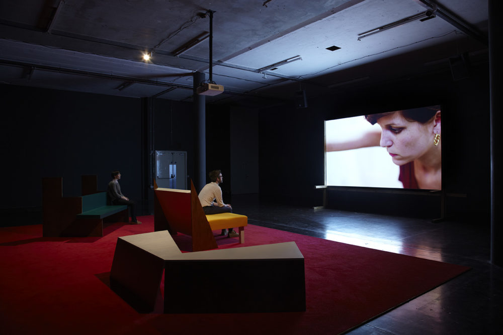 Installation view of Cara Tolmie, Pley (2013). Two visitors sit on angular furniture, watching a film. The screen currently shows a close up of a woman.
