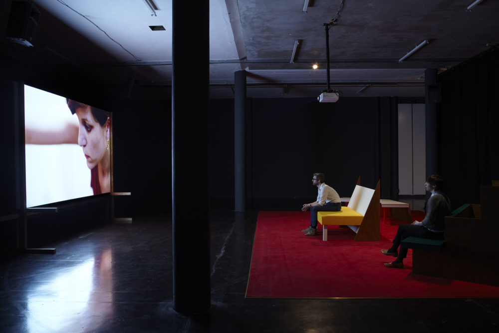 Installation view of Cara Tolmie, Pley (2013). Two visitors sit on angular furniture, watching a film. The screen currently shows a close up of a woman.