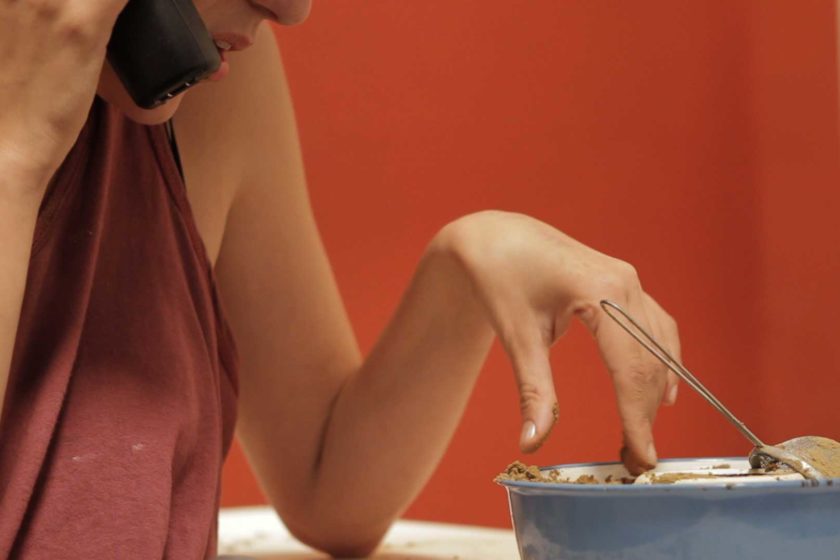 Film still from Cara Tolmie, Pley (2013). A woman is making a phonecall and is sat next to a bowl that contains sand and a tea strainer.