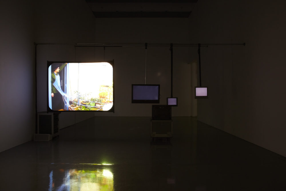 Installation view of Cezary Bodzianowski, Tea Back (2011). A dark room shows five screens, one is illuminated with a projection of a woman stood at her back door.