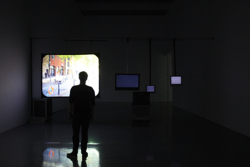 Installation view of Cezary Bodzianowski, Tea Back (2011). A visitor stands in a dark room, looking at a projection of Bristol Harbourside.
