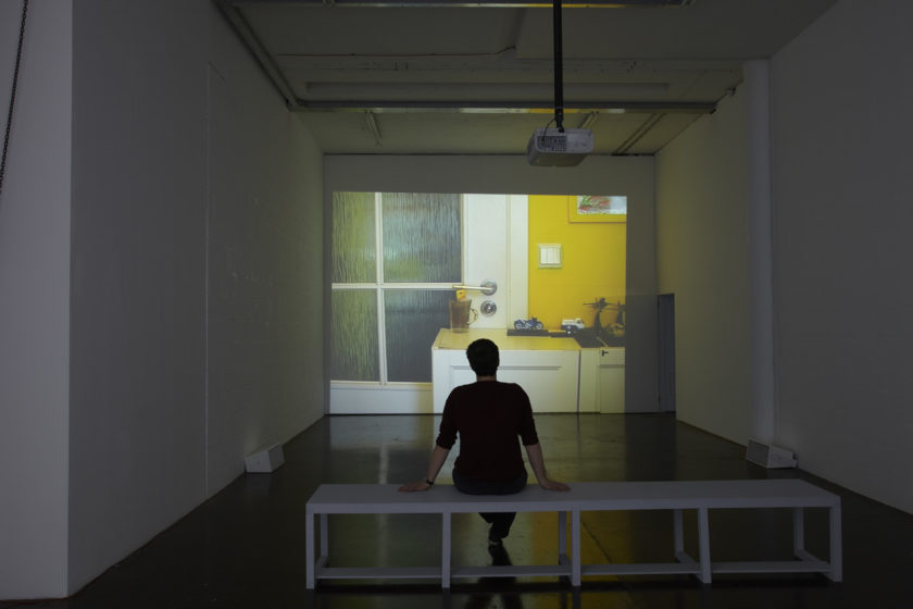 Installation view of Cezary Bodzianowski, Tea Back (2011). A visitor sits on a bench in a room, looking at a projection of a domestic scene.