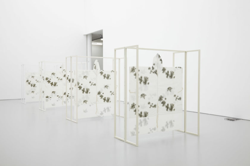 Four white wooden free standing structures stand in the middle of the white gallery. White fabric decorated with dark patches is stretched onto the frames.