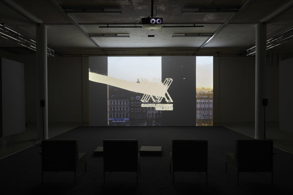 Four large chairs sit on a carpet square in the middle of the gallery. They face a screen that is currently showing a scene of a busy street with an XX logo showing over the image.