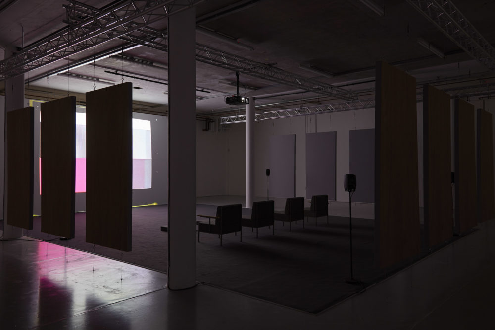 Four large chairs sit on a carpet square in the middle of the gallery. They face a screen that is obscured in this shot by some soundproofing boards that are hanging from the ceiling and framing the seating area.