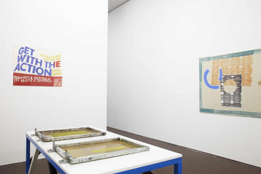 Installation view of Ciara Phillips and Corita Kent, Pull Everything Out (2012) A screen-printing press set up on a table next to screen prints on the wall.