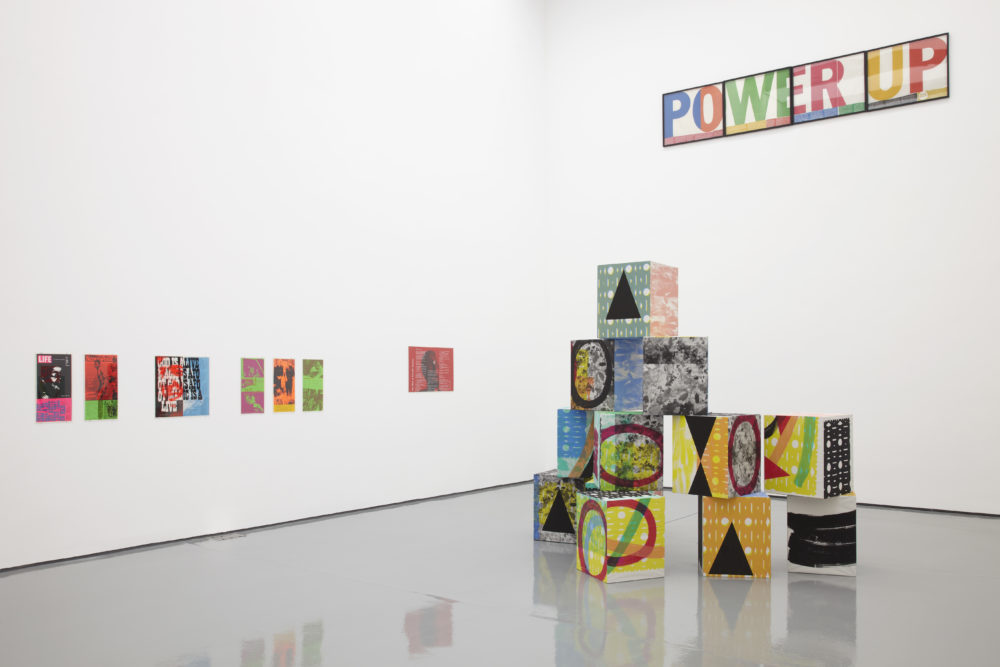 Installation view of Ciara Phillips and Corita Kent, Pull Everything Out (2012) A pyramid of brightly coloured boxes sits in the gallery next to brightly coloured screen prints hanging on the wall.