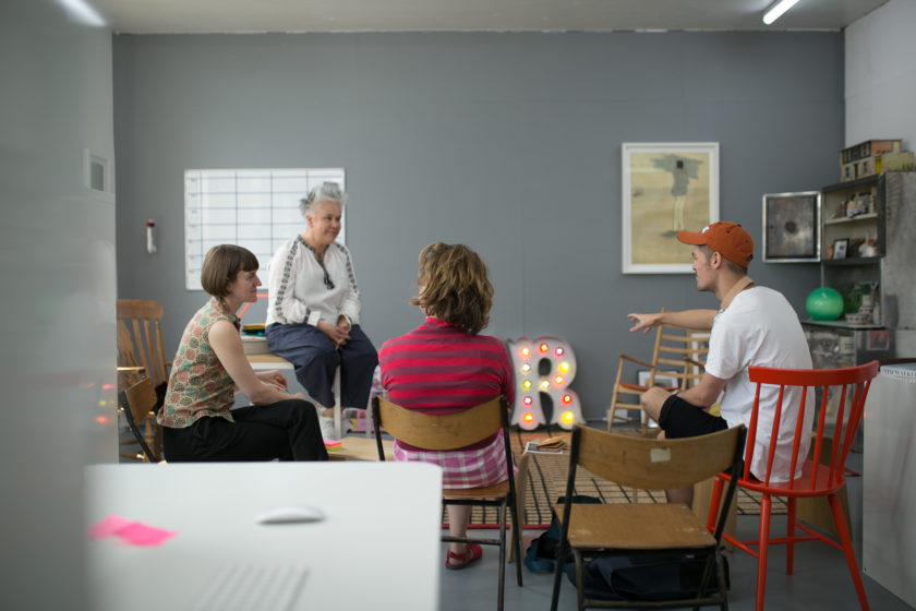 Theatre company Wise Children have a meeting in their studio. Their studio is grey with lots of different types of chairs and a large box light in the shape of the letter R