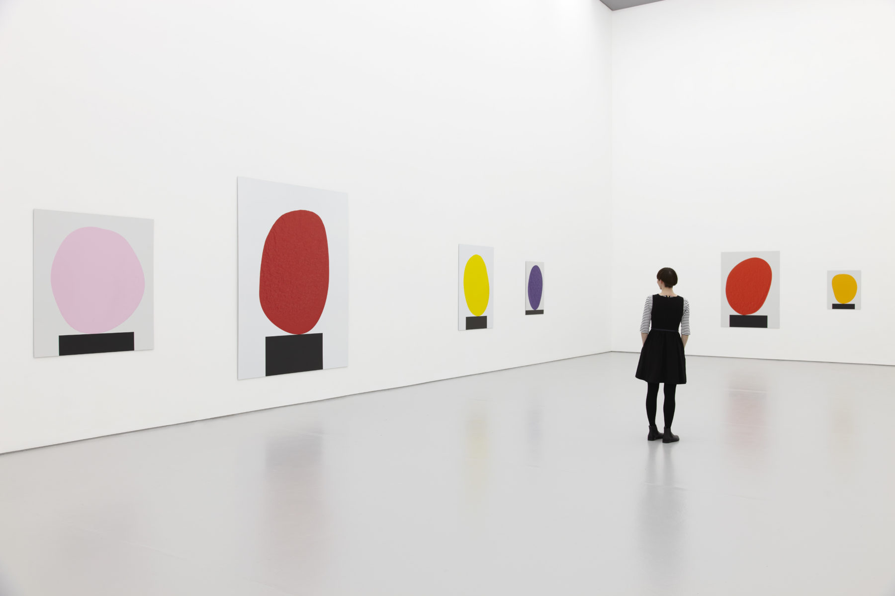 Installation view of David Batchelor Flatlands (2013). A visitor looks at images of coloured blobs balancing on black rectangles.