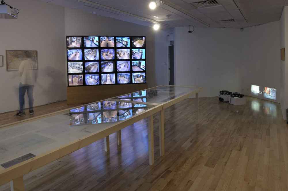 Installation shot: A visitor studies a map, next to a multiscreen installation seemingly showing footage of a gallery. Two glass topped tables in the foreground have works encased.