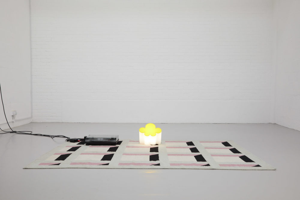 A rug, decorated to look like a building facade with lots of windows lies in the gallery. On top of it is a light in the shape of a cloud and a vinyl record player.