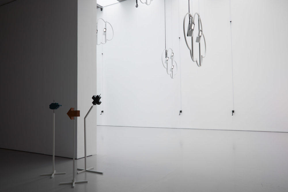 Ceramic way finding sculptures stand at the front of the gallery, suggesting different directions. Behind are cloud shaped metal structures hanging from the ceiling.